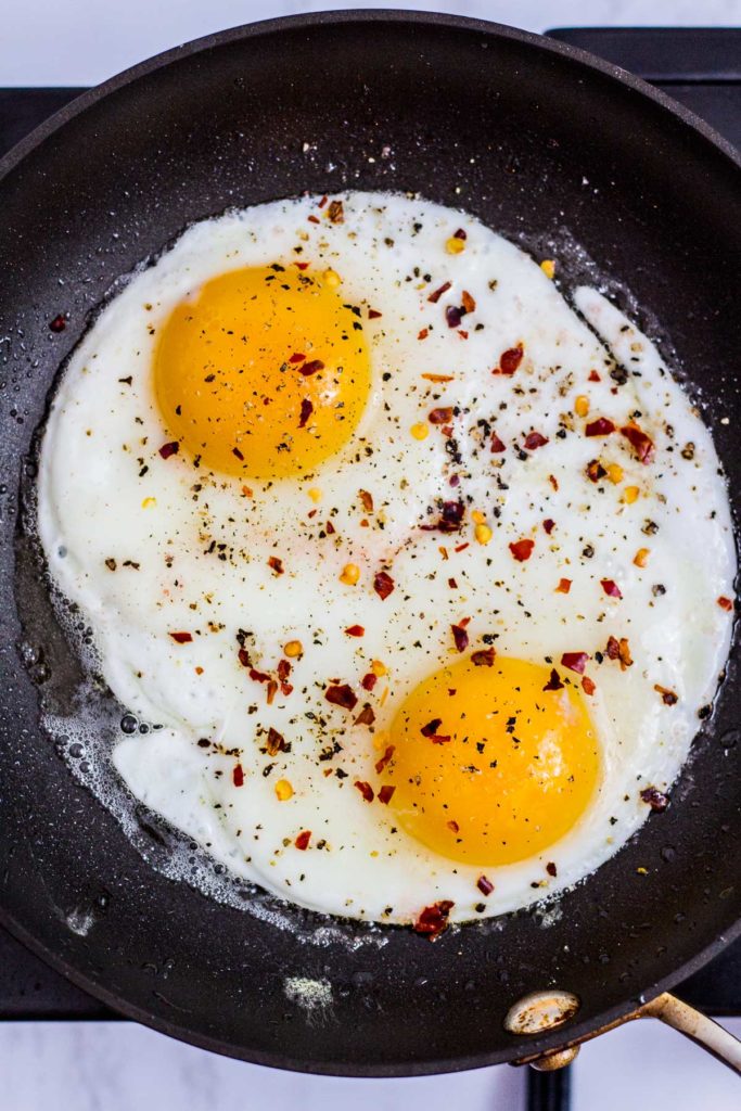 Two fried eggs in the pan seasoned with salt, pepper, and red pepper flakes