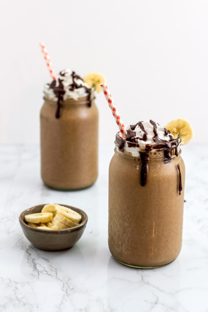 two full mocha flavor banana protein smoothies decorated with coconut whip cream with chocolate syrup drizzle and garnish with banana slice