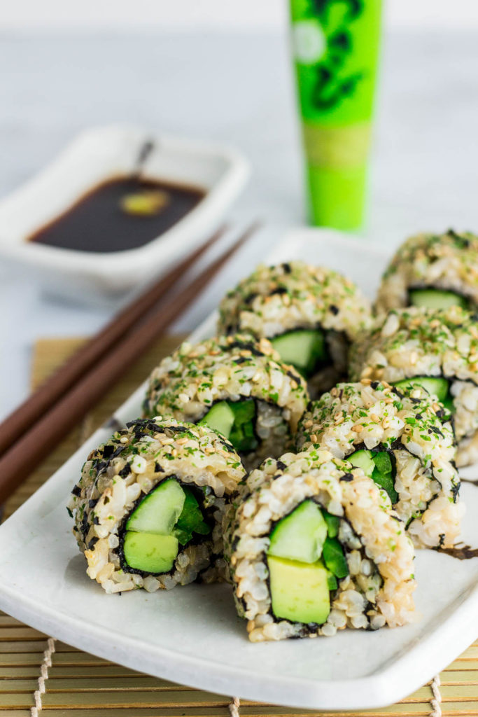 Close up shot of inside out vegan green sushi roll (vegan uramaki). The rolls are placed on top of rectangular plate with soy sauce in the back ground.