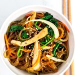 Overhead shot of japchae in a white bowl with chopsticks next to it