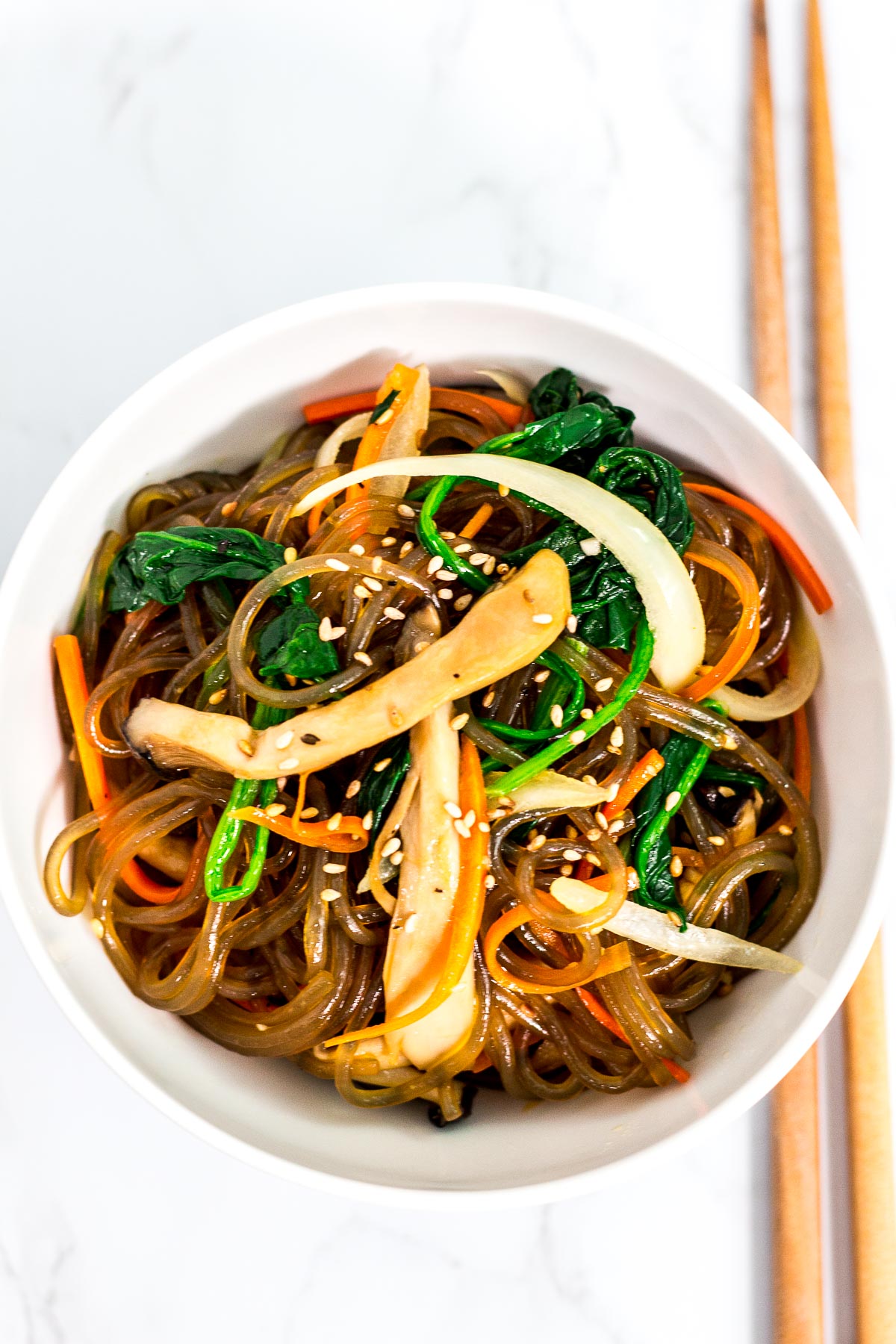 Overhead shot of japchae in a white bowl with chopsticks next to it