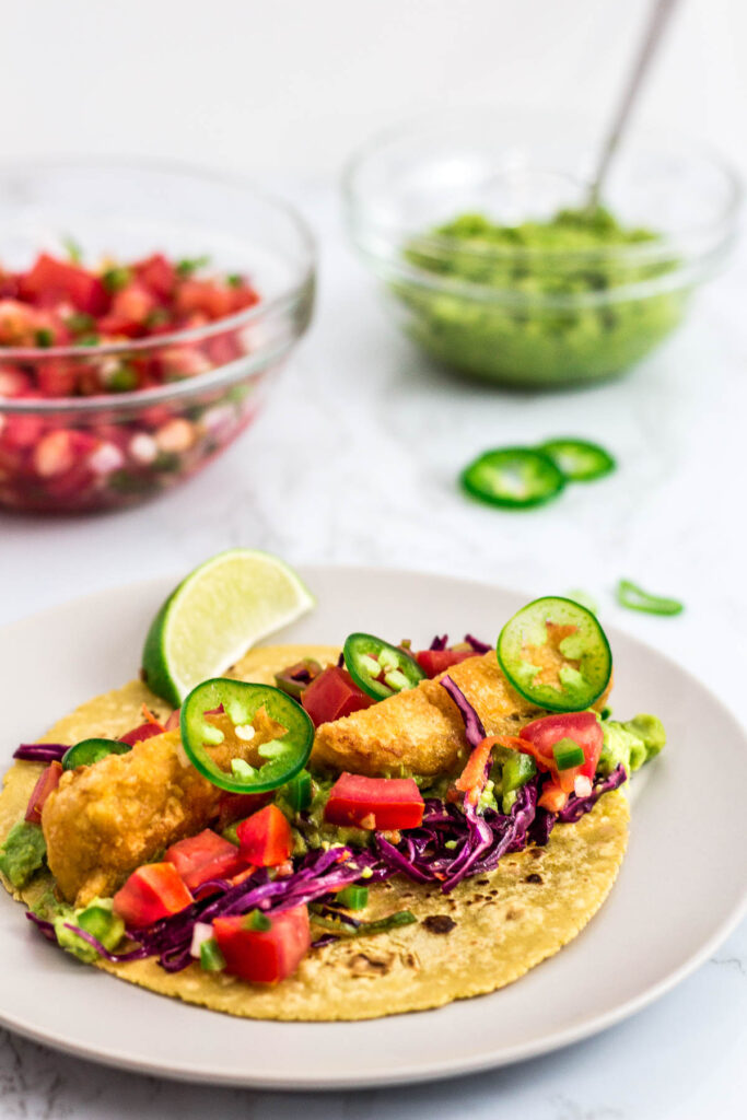 a vegetarian/vegan fish stick tacos on a plate with salsa and guacamole in the background