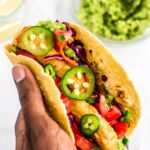 holding a prepared vegan fish stick taco in hand. Guacamole and lime wedges in the background