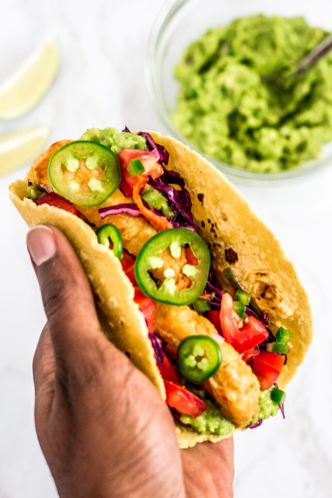 holding a prepared vegan fish stick taco in hand. Guacamole and lime wedges in the background