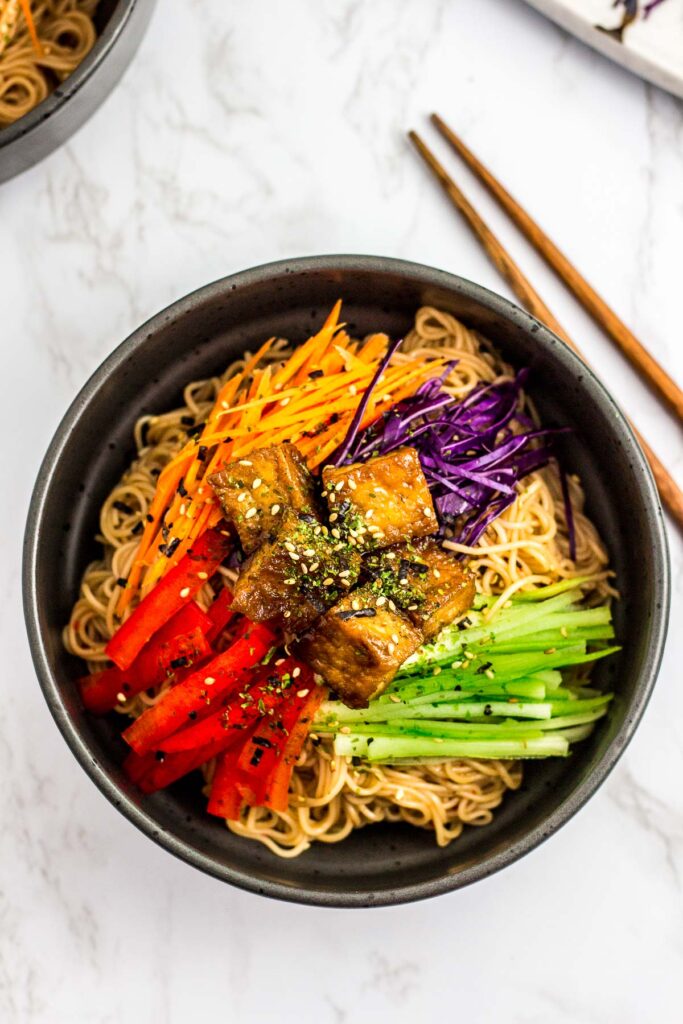 noodle salad in the black bowl with different vegetables and teriyaki tofu on top