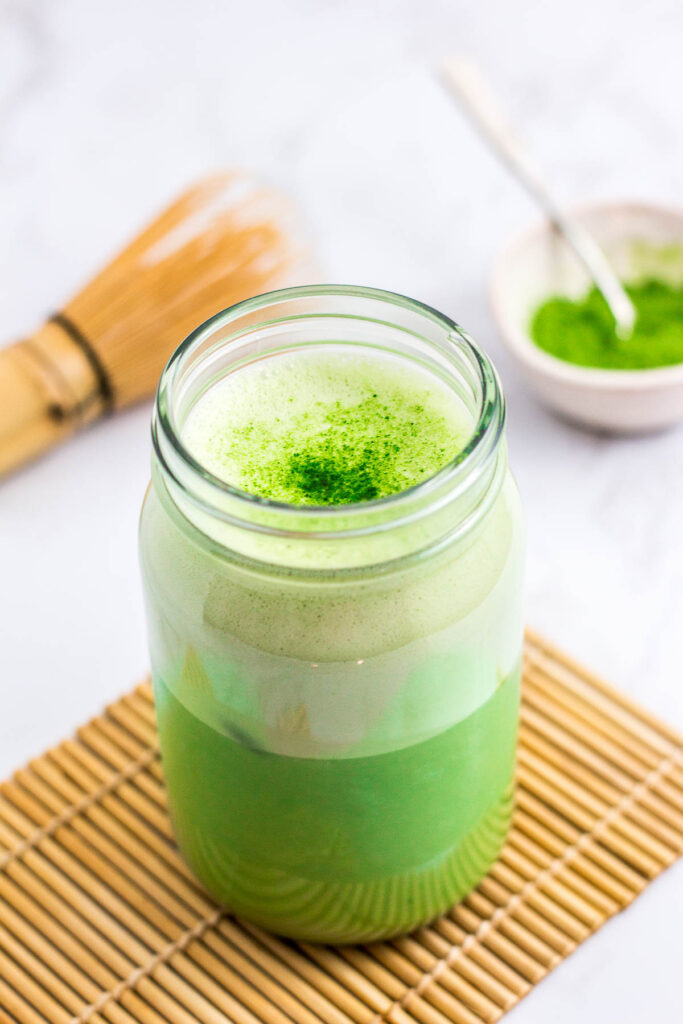 iced matcha latte in a mason jar with matcha powder and whisk in the background