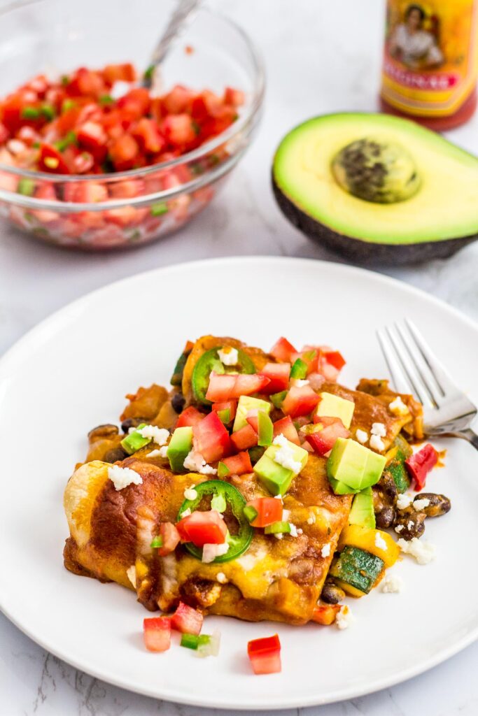 summer veggie enchiladas on a white plate with salsa, avocado and hot sauce in the back ground