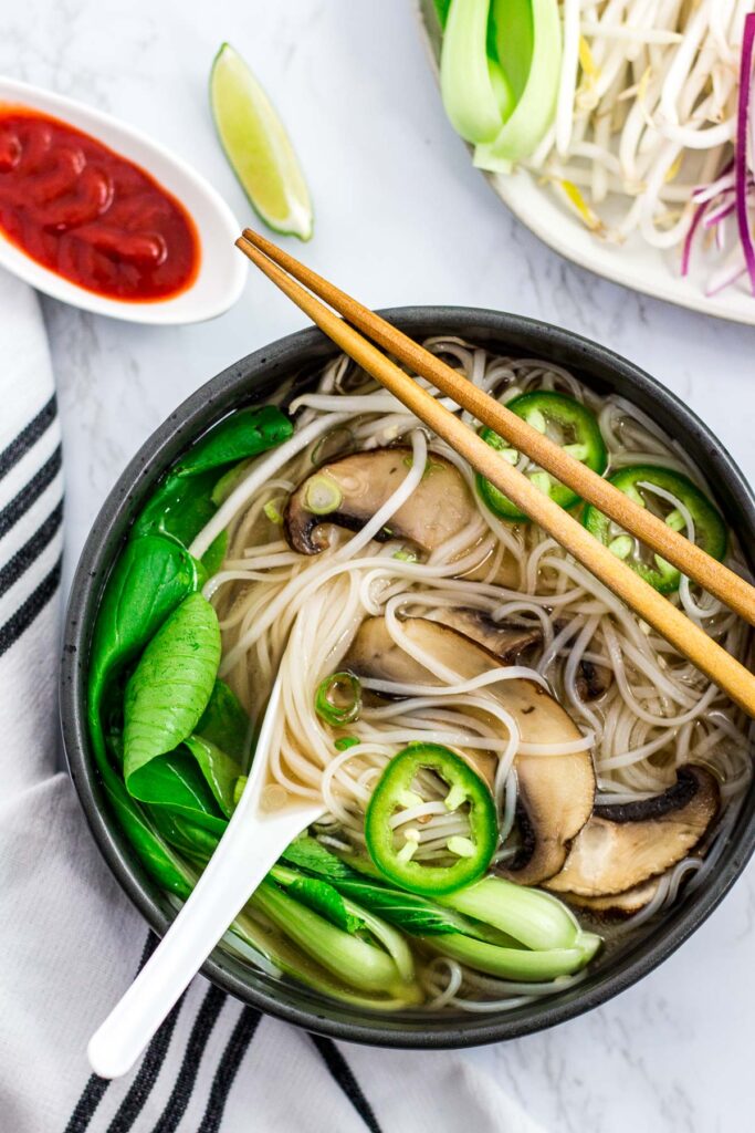 Vegan pho with mushroom, bok choy, and jalapeno in a large black bowl with sriracha sauce on the side