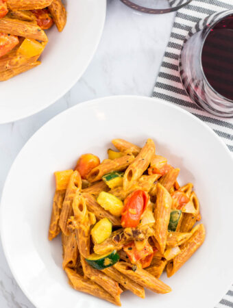 two bowls of vegan creamy lentil pasta primavera with a glass of red wine in the background