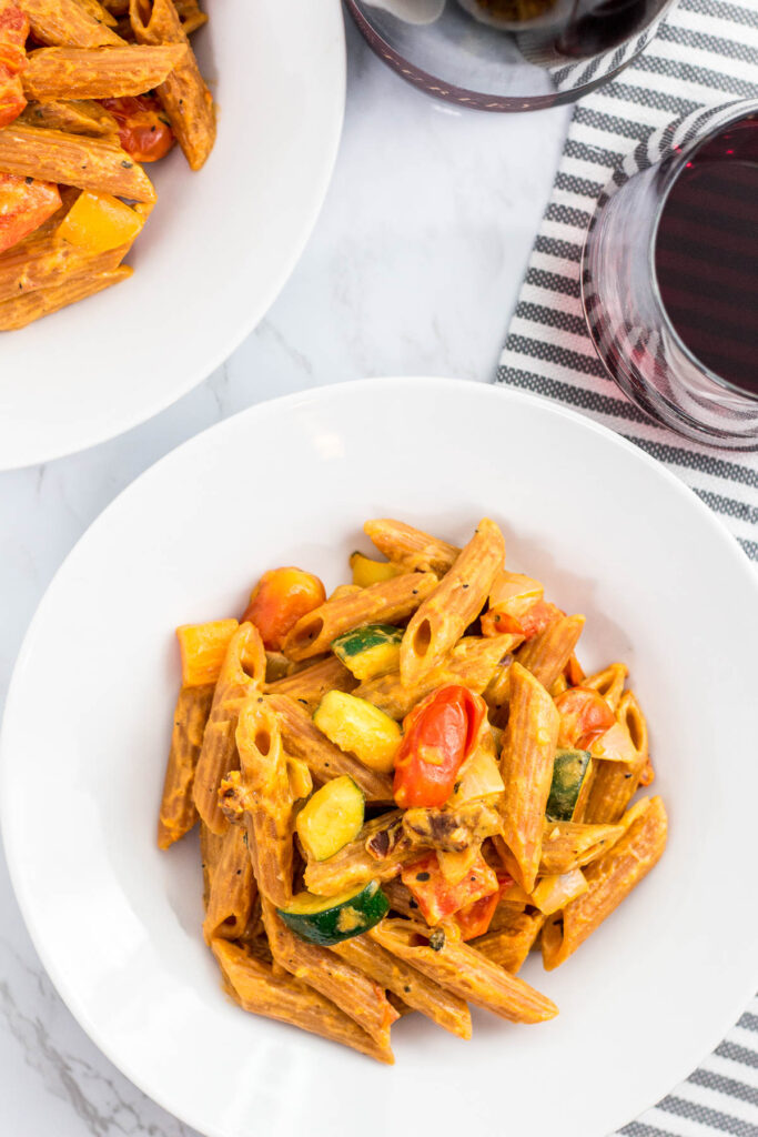 two bowls of vegan creamy lentil pasta primavera with a glass of red wine in the background