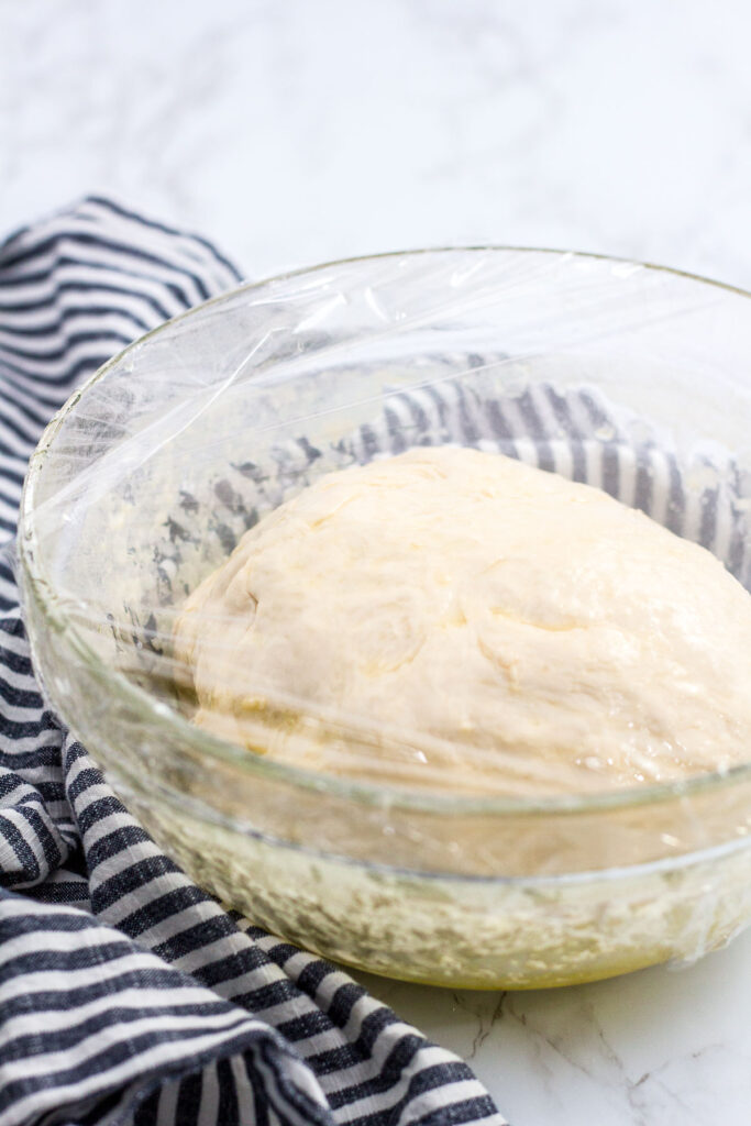 Pizza dough in a clear bowl, covered with plastic wrap