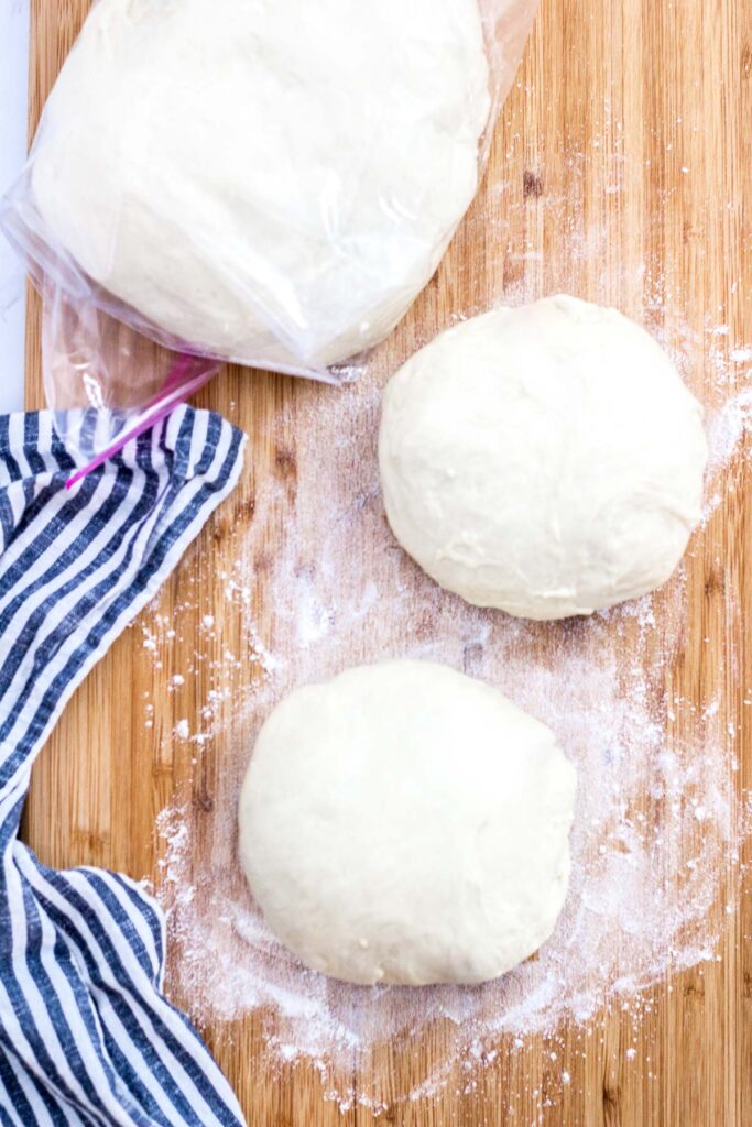 overhead shot of one pizza dough in a freezer bag, and two smaller pizza dough on a cutting board