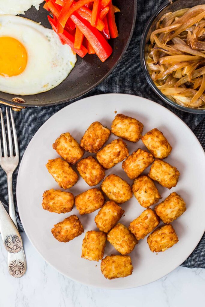 air fried tater tots on a plate, caramelized onion, sauteed pepper, and fried egg in the background