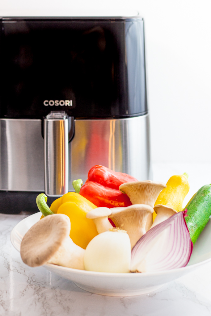 Mushroom, bell pepper, yellow squash, zucchini, and onion in a white bowl in front of Cosori air fryer