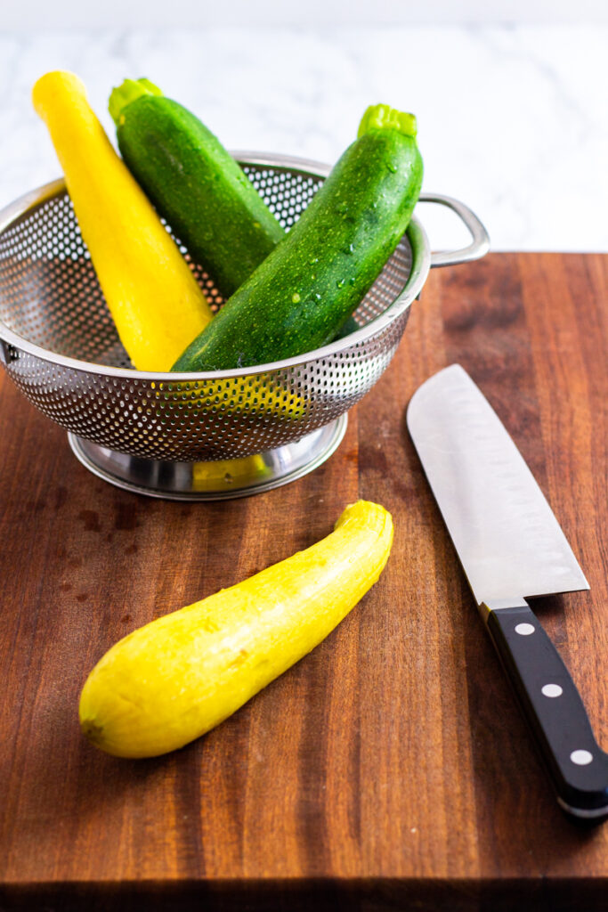 two washed zucchini and one yellow squash in a colander and one yellow squash on a cutting board.