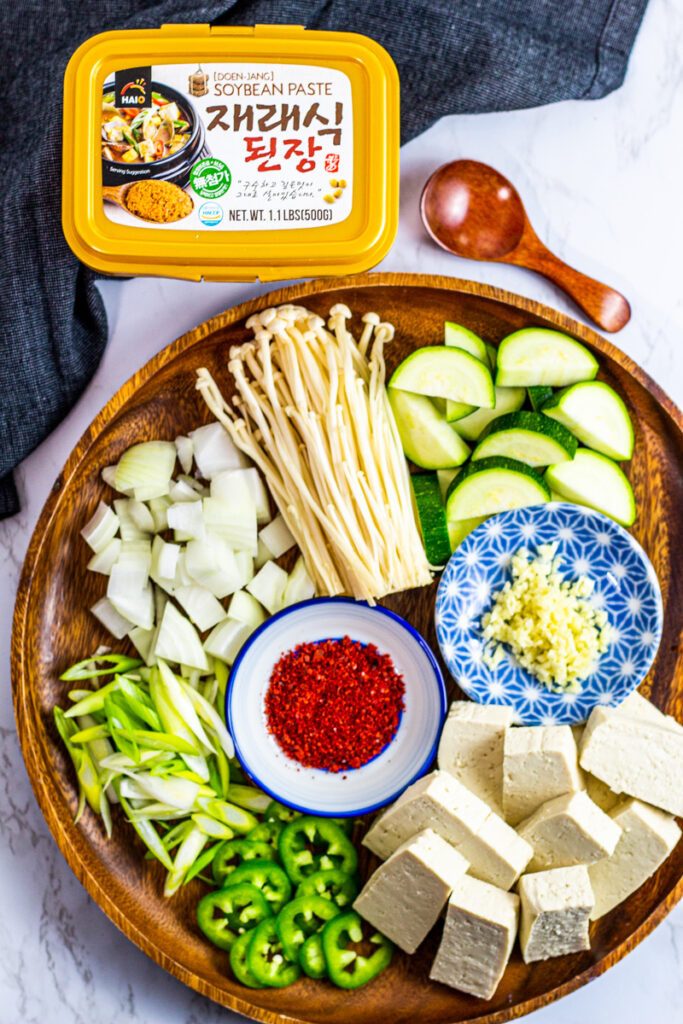 ingredients for the stew - zucchini, minced garlic, tofu, jalapeno, green onion, onion, gochugaru, enoki mushroom, and doenjang.