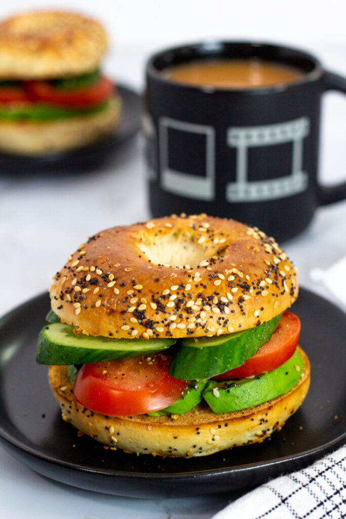 close up photo of vegan avocado bagel with tomato and cucumber with a cup of coffee in the background.