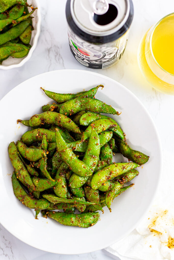 spicy edamame on a white plate with a glass of beer and the can next to it.