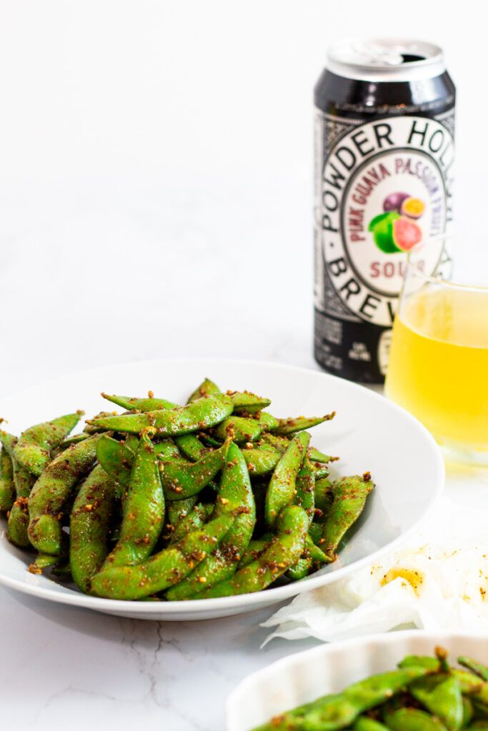 prepared edamame on a white plate with a glass of beer next to it.