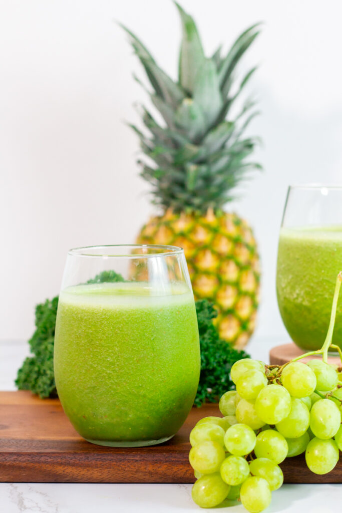 close up shot of green smoothie with kale, pineapple, and grape in the background.