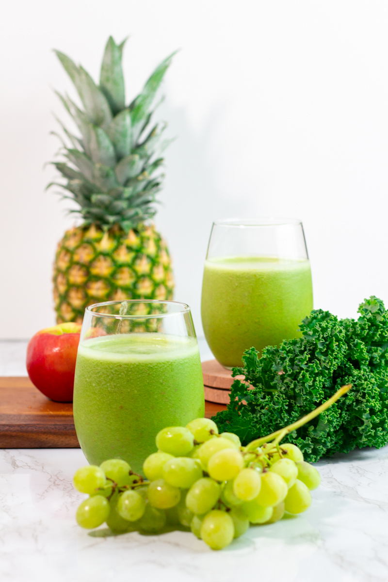 two glasses of kale pineapple green smoothie with kale and fruits in the background.
