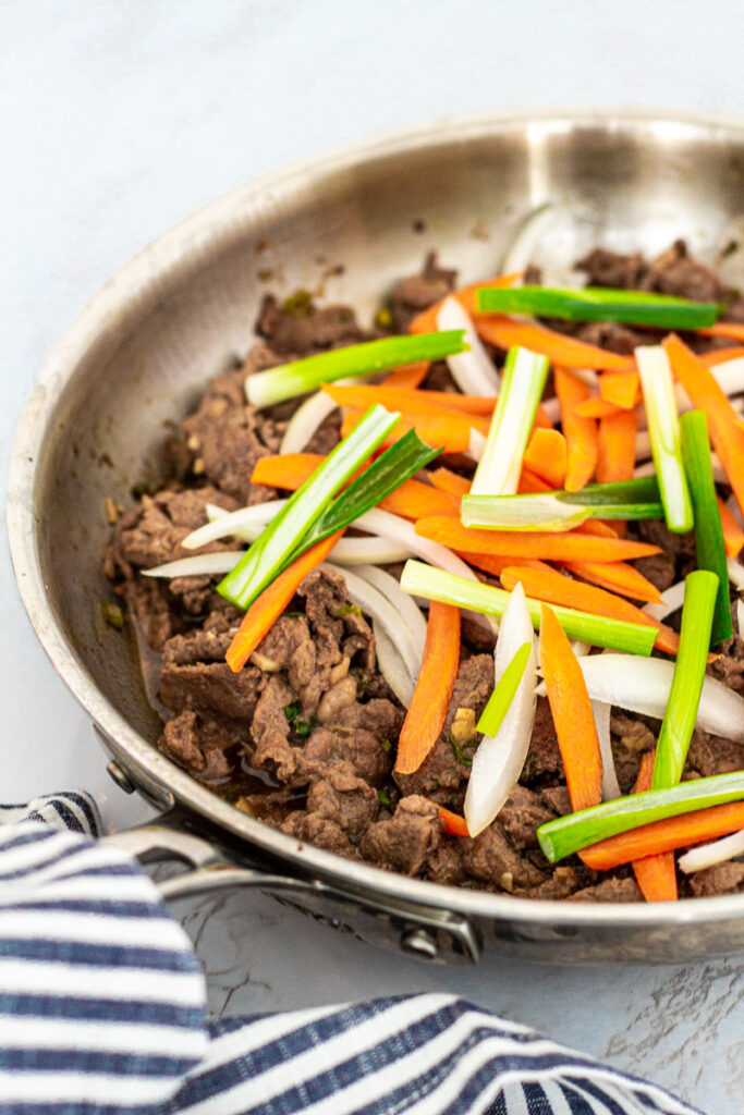 Cooked beef in the pan topped with fresh onion, green onion, and carrot on top
