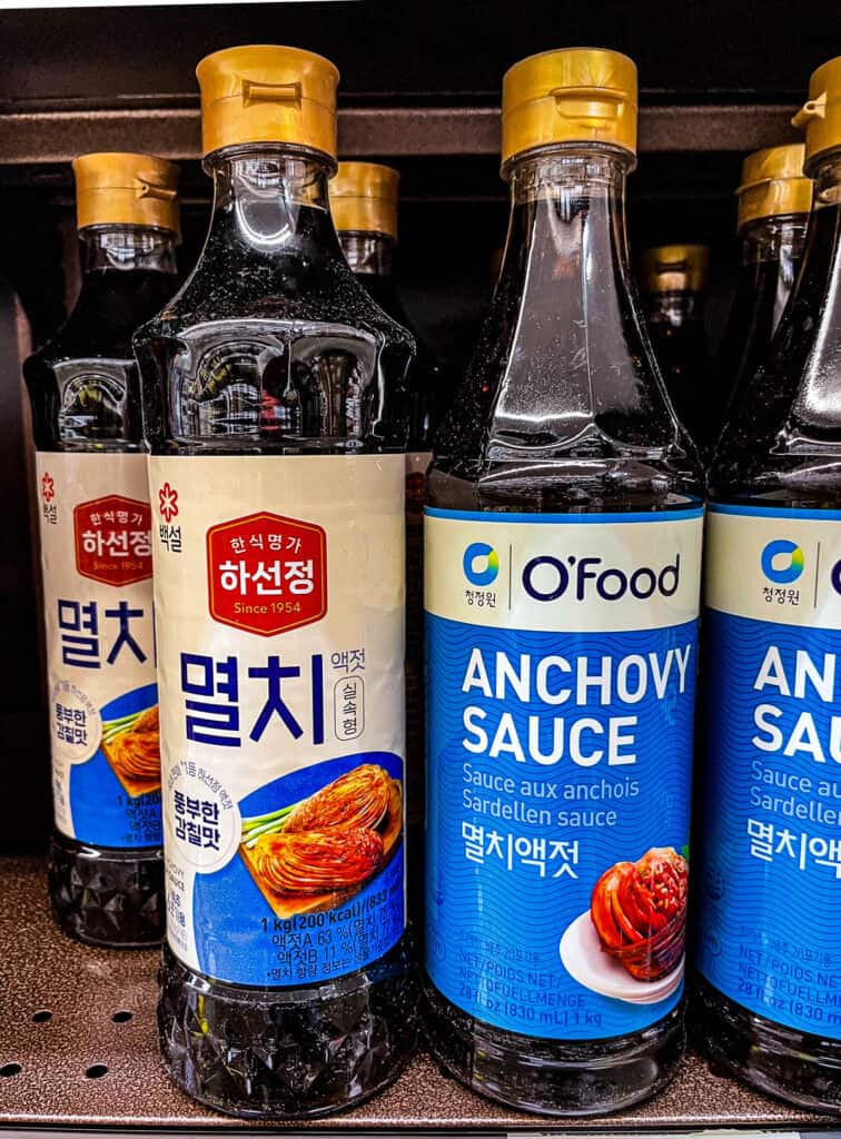 Close-up of Korean anchovy fish sauce bottles, including Baeksul and O'Food brands on a grocery shelf