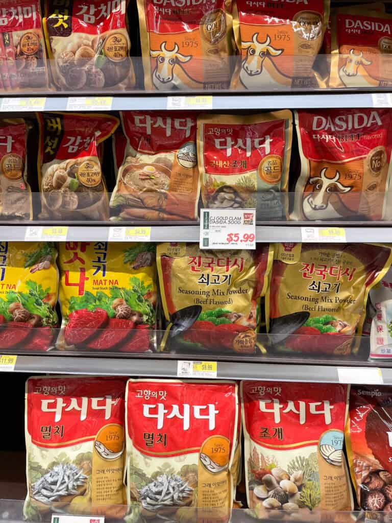 A grocery shelf featuring a variety of Korean soup stock powders including beef, anchovy, clam, and mushroom flavors, with popular brands like Dasida and Matna on display