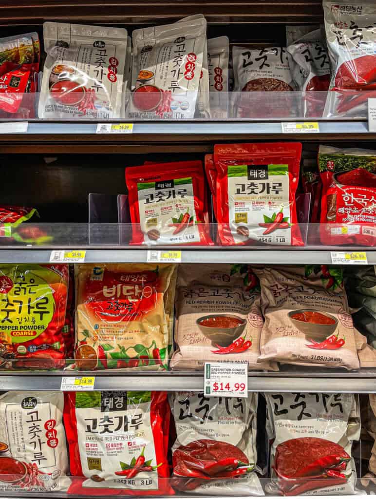 A grocery store shelf displaying multiple brands and sizes of Korean gochugaru, including coarse and fine types used for kimchi, soups, and marinades