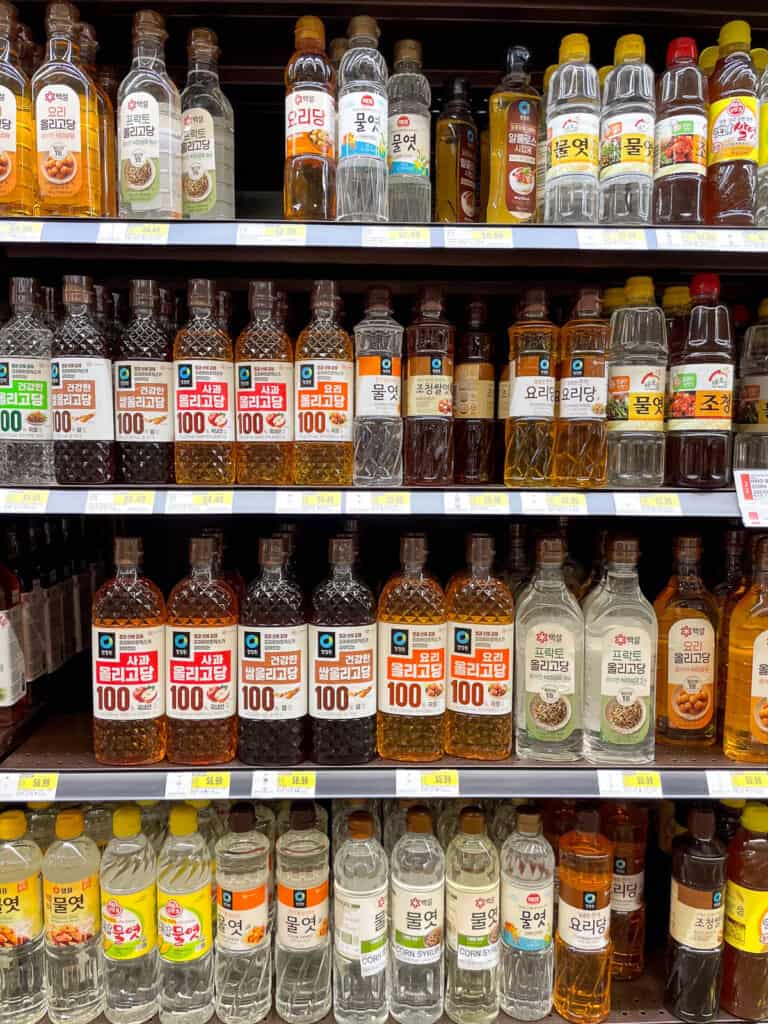 A variety of Korean corn syrup, rice syrup, and cooking syrup brands displayed on multiple shelves at an Asian grocery store