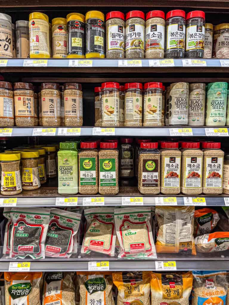 A wide selection of Korean sesame seed products, including roasted, unhulled, and powdered sesame seeds in jars and bags on a grocery store shelf