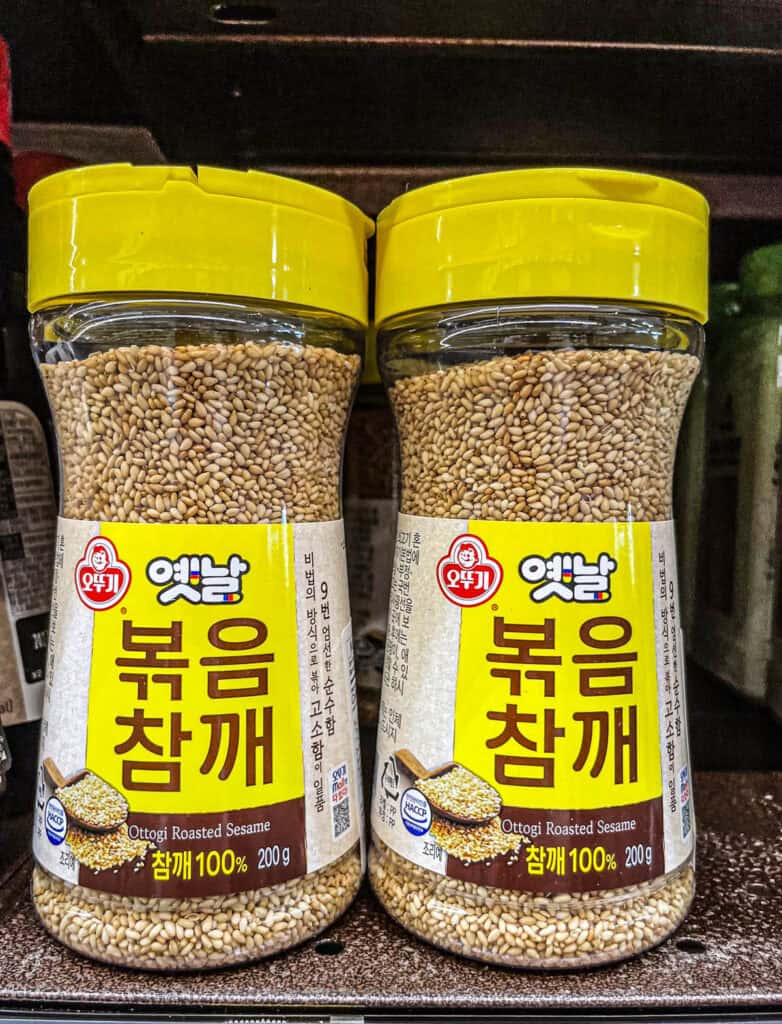 Close-up of two containers of Ottogi brand roasted sesame seeds with yellow caps, labeled in Korean and English