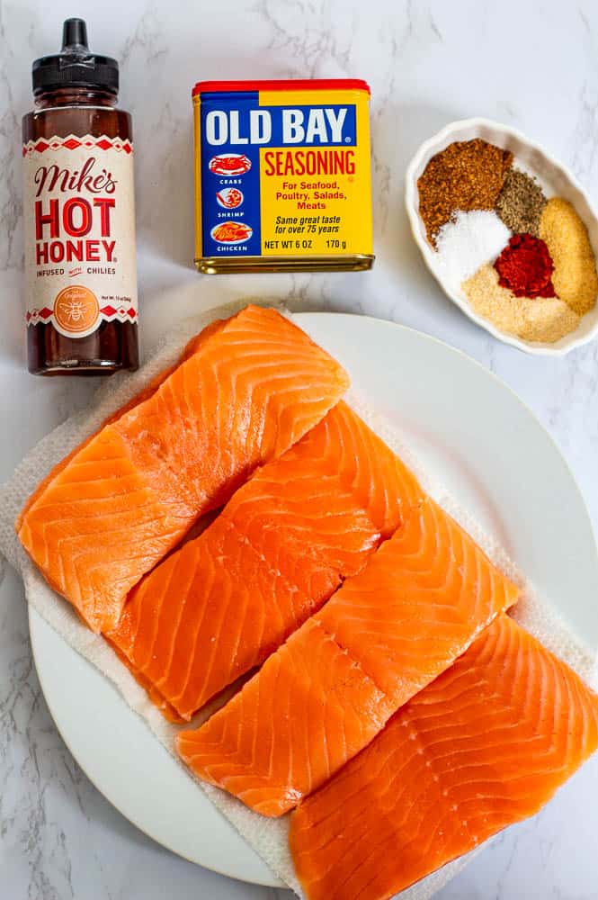 Raw salmon fillets on a plate with Old Bay seasoning, Mike's Hot Honey, and a dish of spices