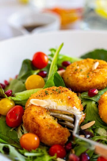 Close-up of crispy fried goat cheese on a bed of salad greens, sliced with a fork, surrounded by cherry tomatoes and pomegranate seeds