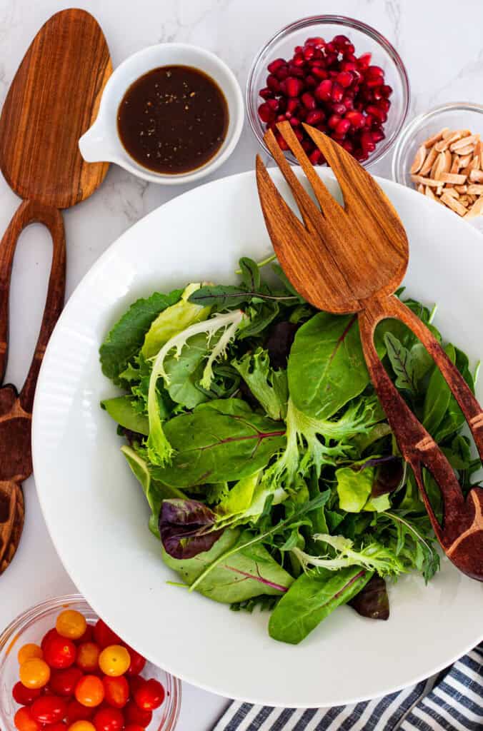 Overhead view of a white bowl filled with spring mix greens, surrounded by pomegranate seeds, cherry tomatoes, toasted almonds, and balsamic dressing with wooden salad servers