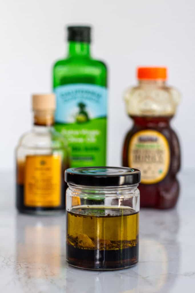 Jar of homemade balsamic honey vinaigrette with olive oil, balsamic vinegar, Dijon mustard, and honey bottles blurred in the background