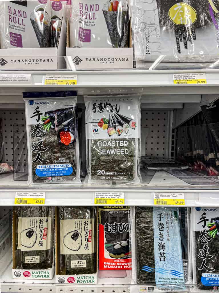 Packages of roasted seaweed for hand rolls and onigiri rice balls displayed on a Japanese grocery store shelf, showing different brands and cuts.