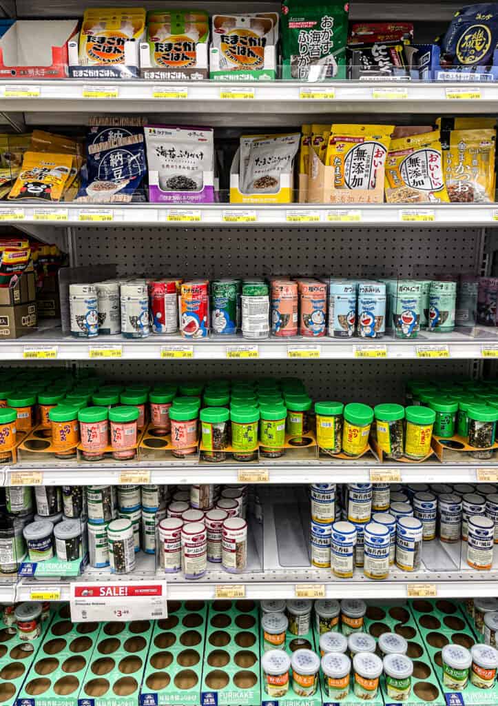 Assorted jars and packets of furikake rice seasoning in various flavors displayed on a Japanese grocery store shelf.