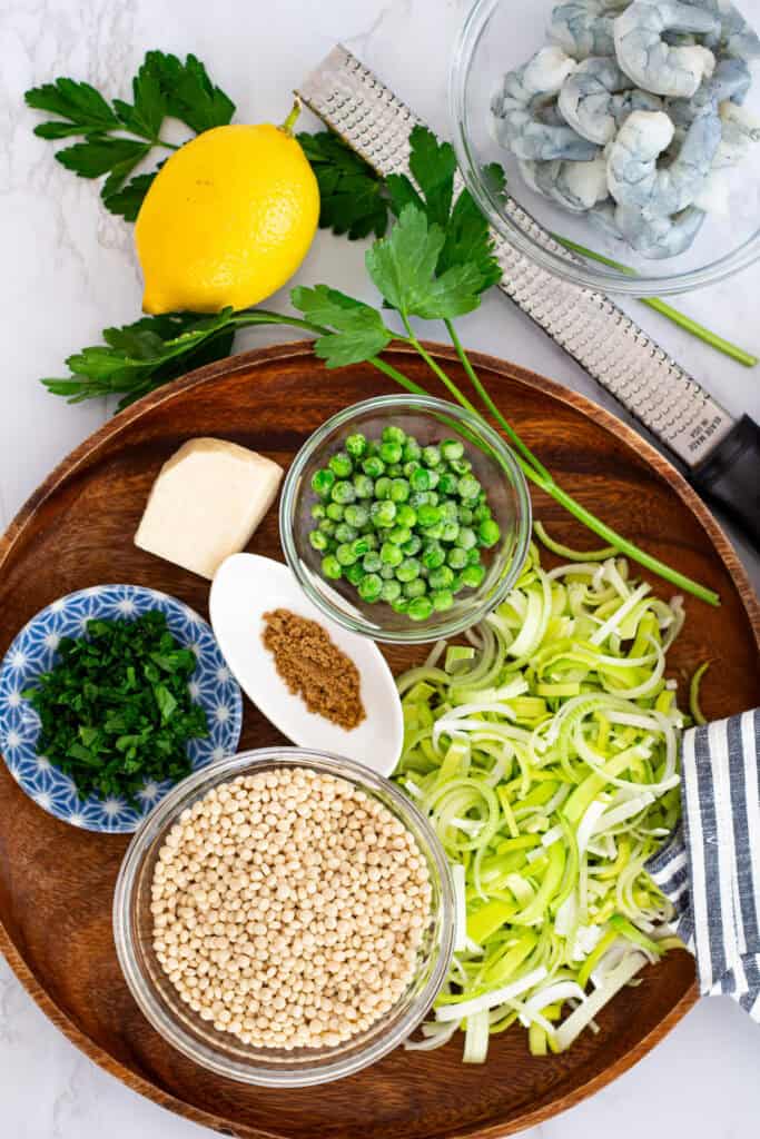 Flat lay of ingredients for couscous risotto: Israeli couscous, shrimp, leeks, parsley, lemon, Parmesan, and peas