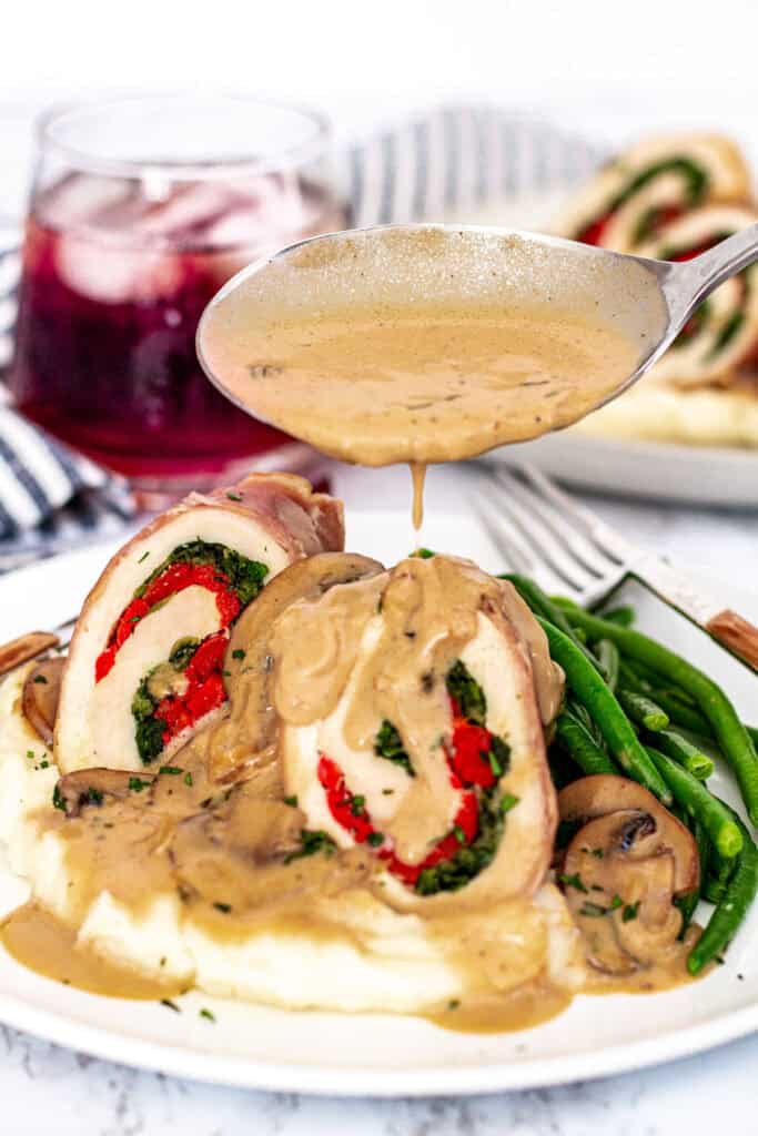 Spoon drizzling creamy mushroom marsala sauce over chicken roulade with mashed potatoes and green beans.