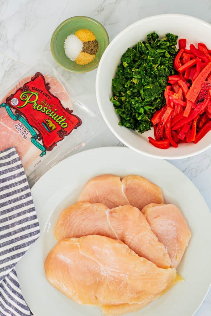 Ingredients for chicken roulade including chicken breasts, prosciutto, spinach, red peppers, and spices on a counter.