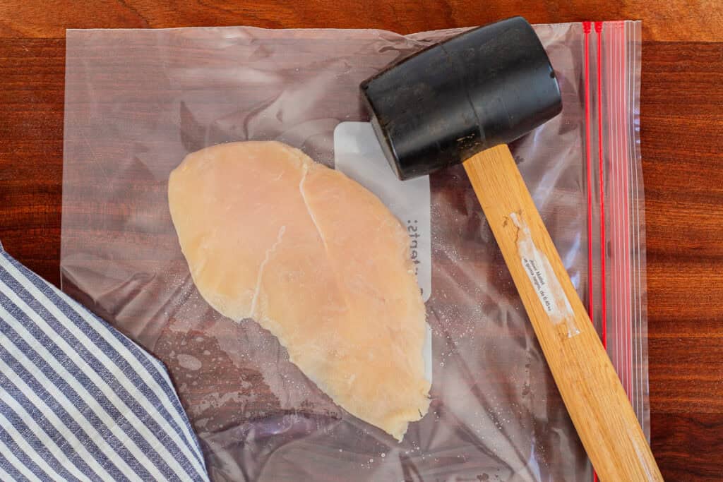 Raw chicken breast placed in a plastic bag with a rubber mallet for chicken roulade.