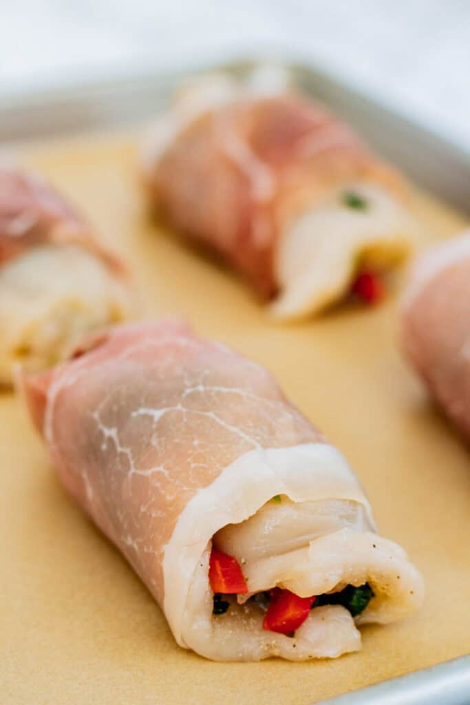 Rolled chicken breast wrapped in prosciutto on a baking sheet.