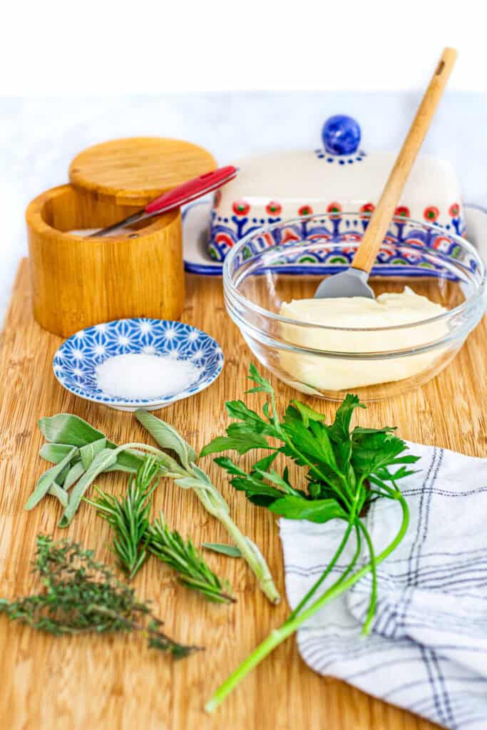 Glass bowl of softened butter with fresh rosemary, thyme, sage, parsley, and kosher salt on a wooden board, ready to make homemade herb butter