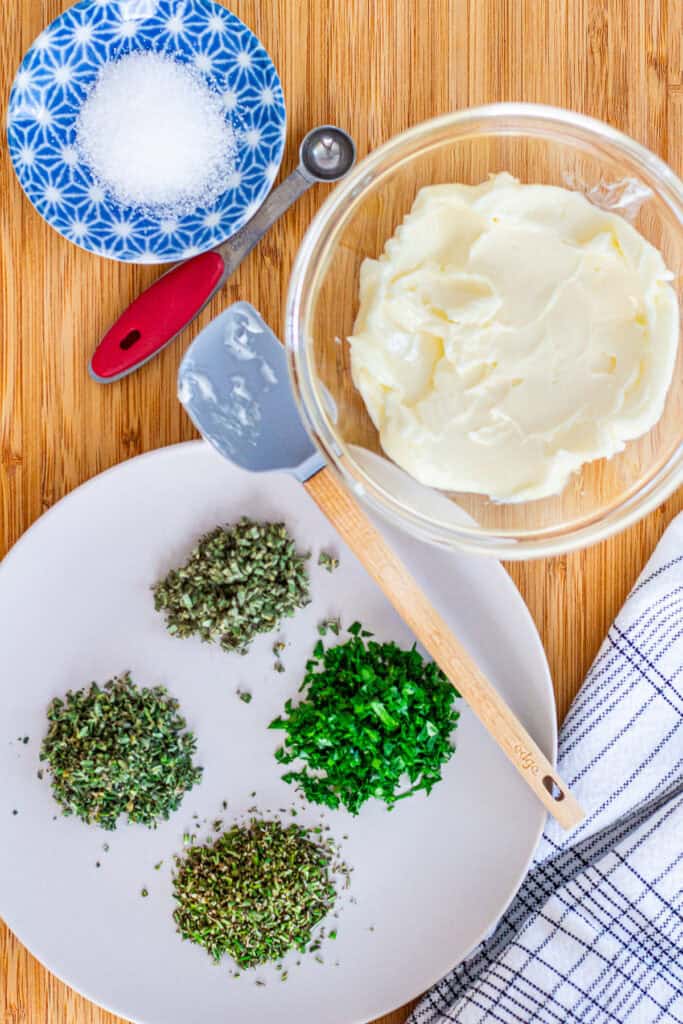 Overhead view of butter, fresh herbs, and kosher salt arranged neatly on a wooden cutting board for making fresh herb butter.