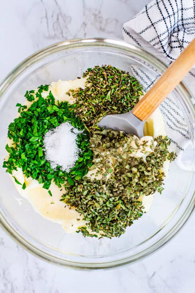 Bowl of softened butter with sprigs of fresh rosemary, thyme, sage, and parsley on a wooden surface for compound butter.