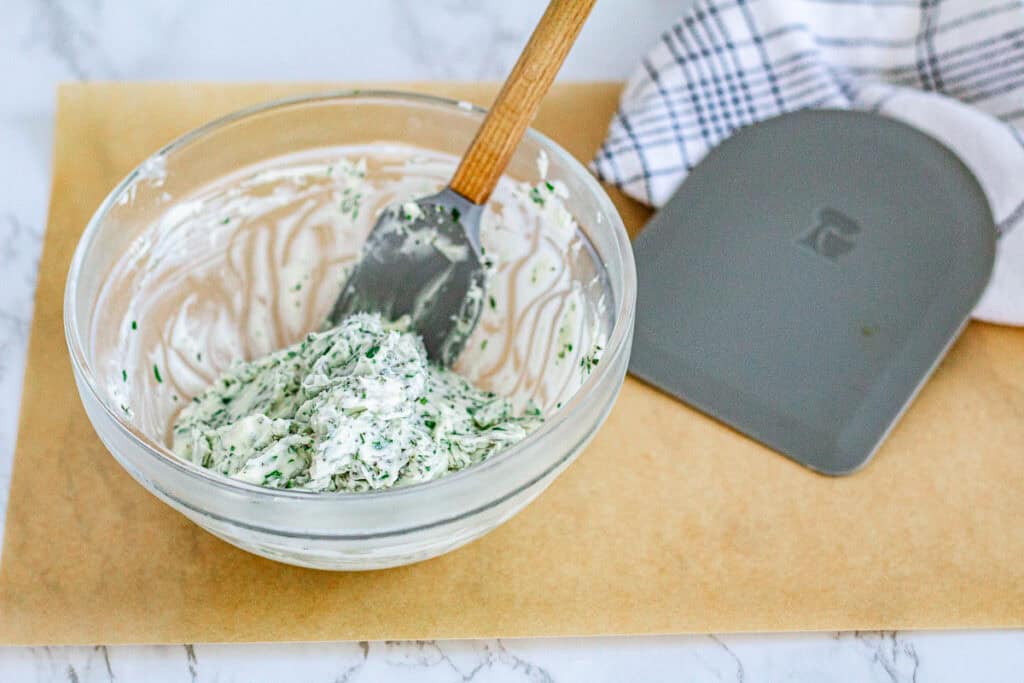 Softened butter mixed with fresh rosemary, thyme, sage, parsley, and kosher salt in a glass bowl for a Thanksgiving herb butter recipe.
