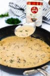 Spoon lifting marsala cream sauce with mushrooms from a skillet, showing creamy texture.