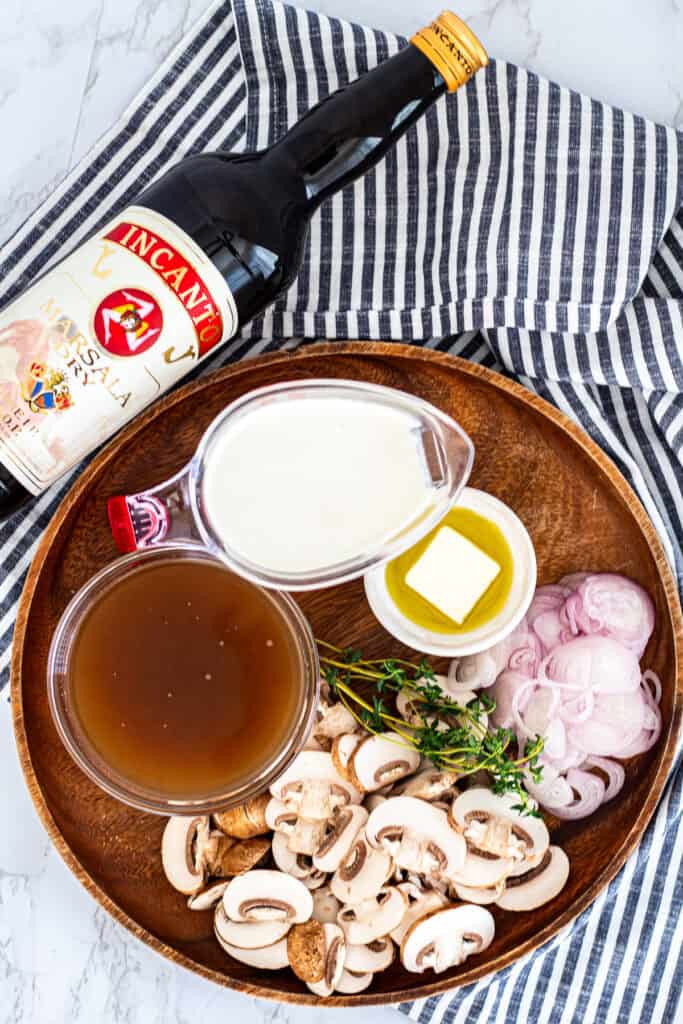 Ingredients for marsala cream sauce on a tray, including Marsala wine, cream, broth, mushrooms, shallots, butter, olive oil, and thyme.