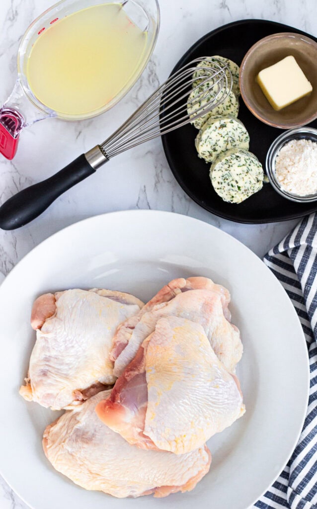 Ingredients for Thanksgiving chicken thighs including raw chicken thighs, herb butter, chicken broth, flour, and butter on a kitchen counter.